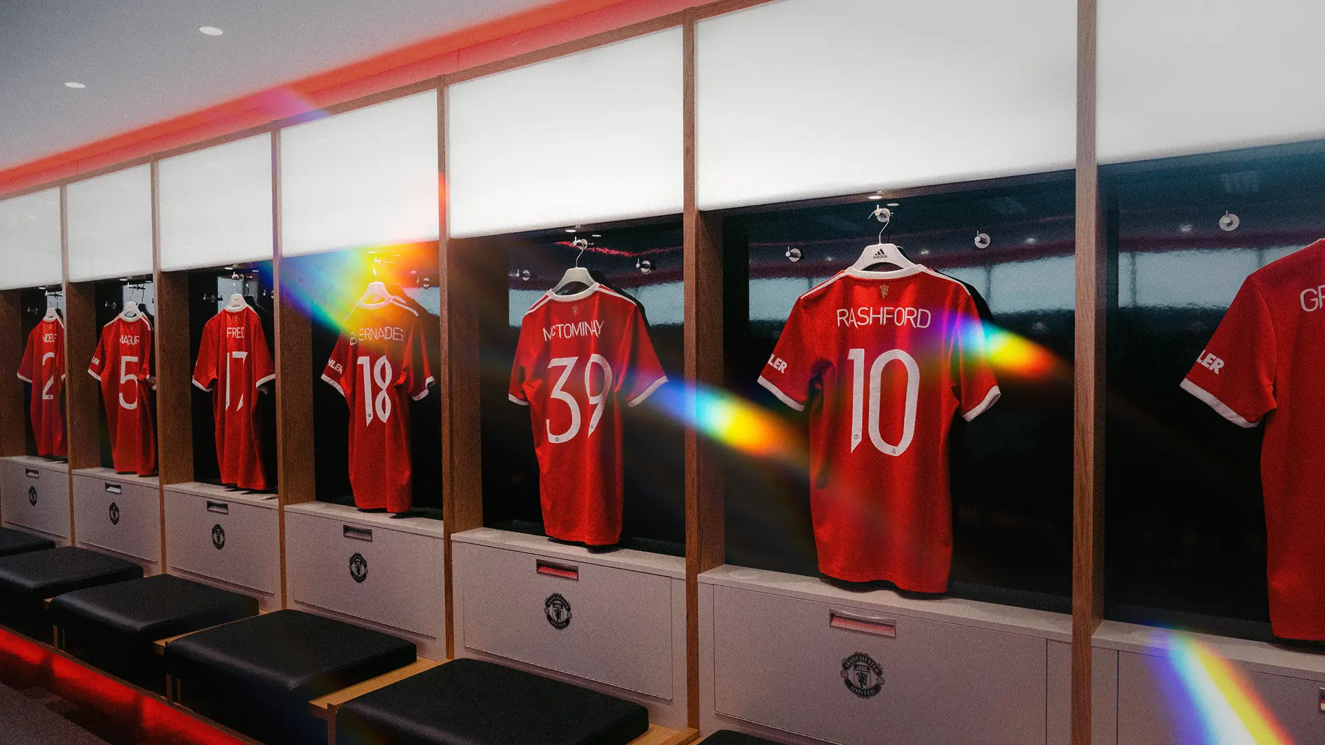 Manchester United home shirts hanging in the Old Trafford dressing room, with Rashford number 10 and McTominay number 39 in view and a light flare across the frame.