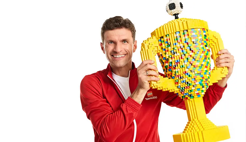 Thomas Muller wearing a red jacket and holding a colourful LEGO trophy, symbolising his collaboration with global brands and family-friendly partnerships that enhance brand image.