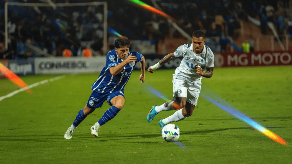 Two football players competing for the ball during a CONMEBOL match at a floodlit stadium, with perimeter advertising boards visible in the background.