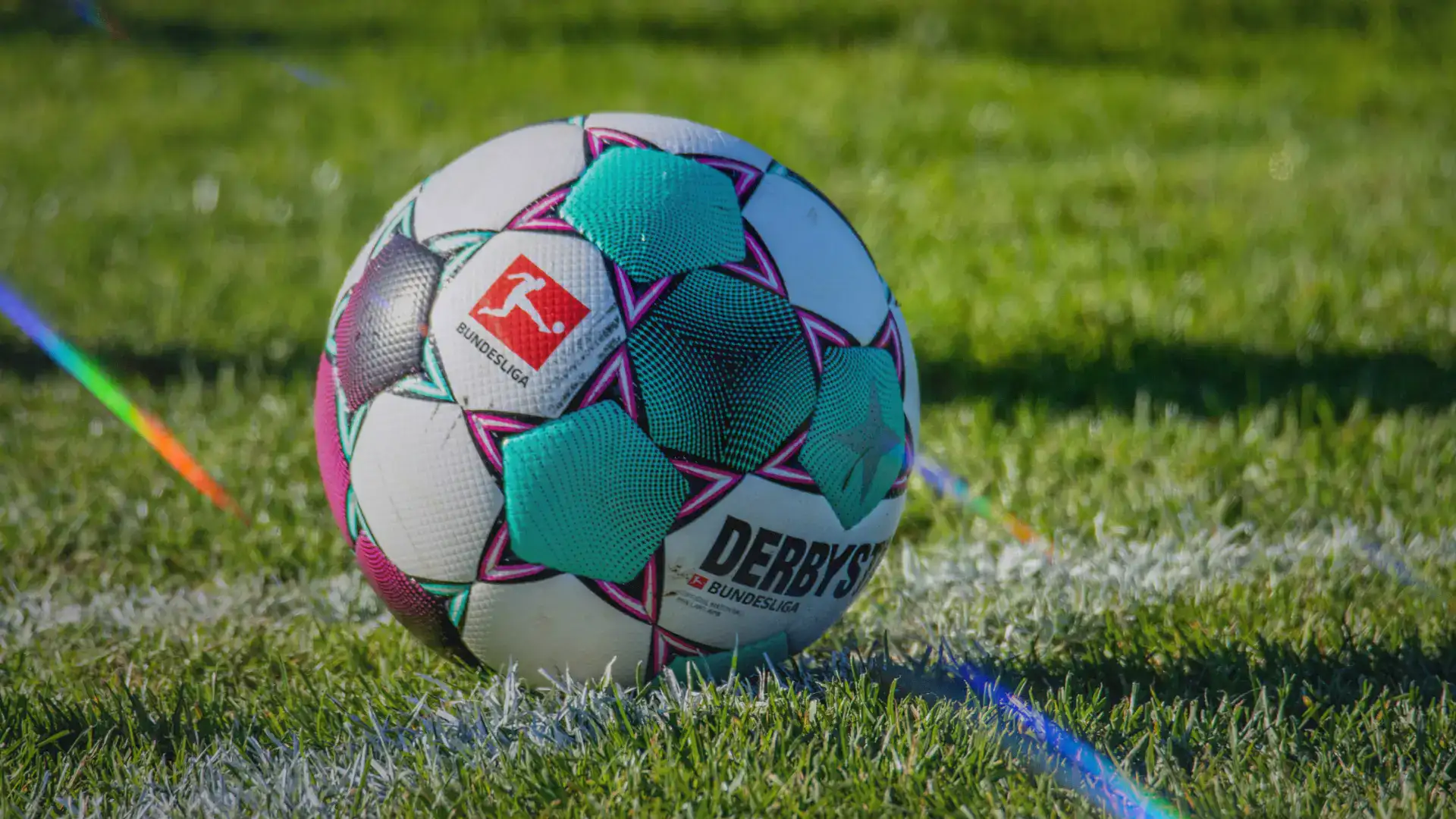 Close-up of a Bundesliga match ball with turquoise and pink patterns resting on green grass near a white line.